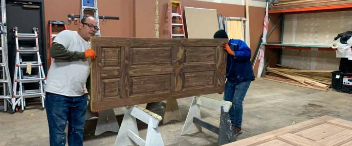 two workers lifting a fabricated door inside their shop, preparing to be painted