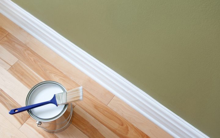 Painting the baseboard, on the floor is a can of white paint and a brush on top of the can